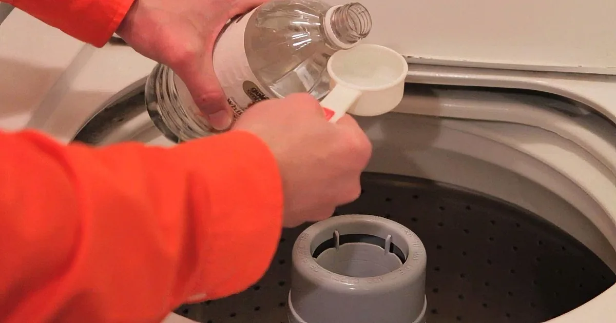A person in an orange shirt pours liquid from a bottle into a measuring cup over a washing machine.