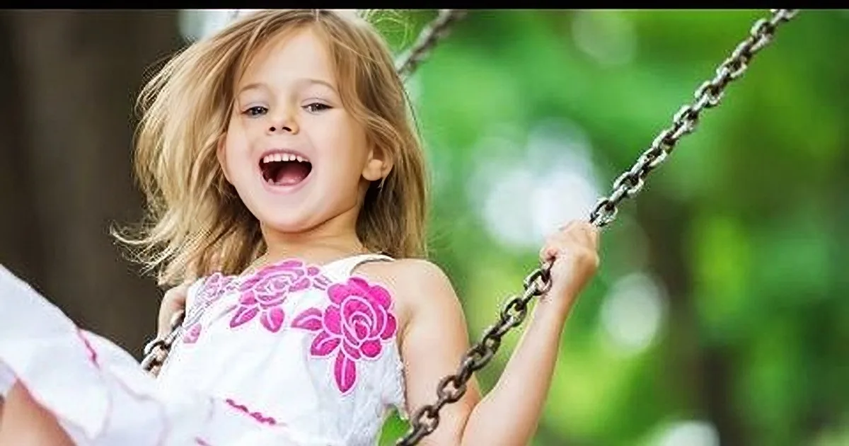 A joyful child swings on a swing set, wearing a pink floral dress, surrounded by greenery.