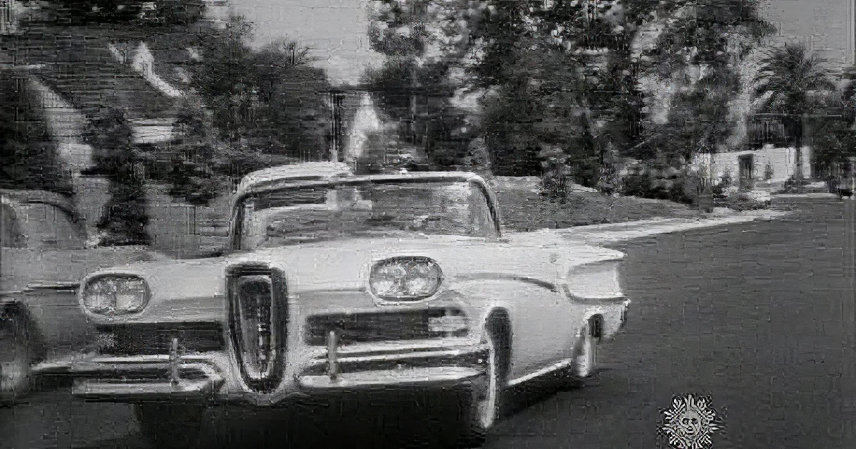A vintage Edsel car is driving on a suburban street with trees and houses in the background.