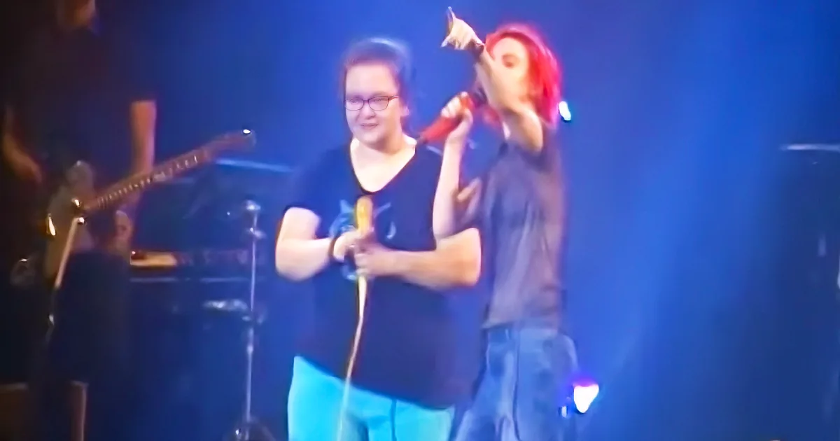 A person with red hair holds a microphone, singing on stage with another person in glasses nearby.