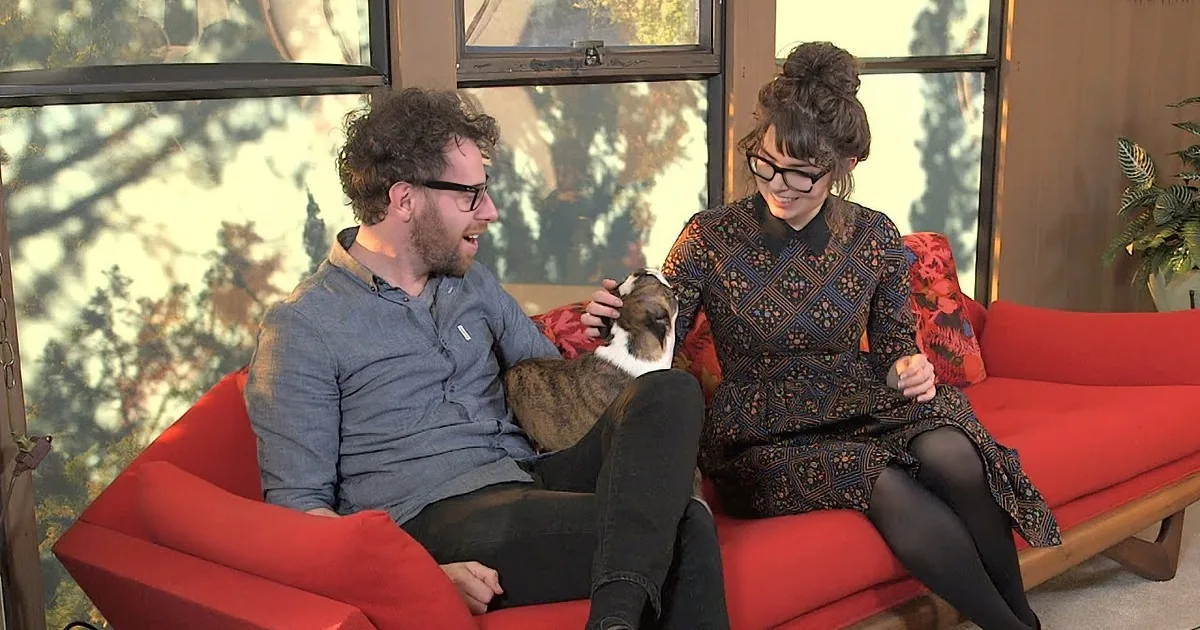 A man and woman sit on a red couch, laughing and petting a dog.