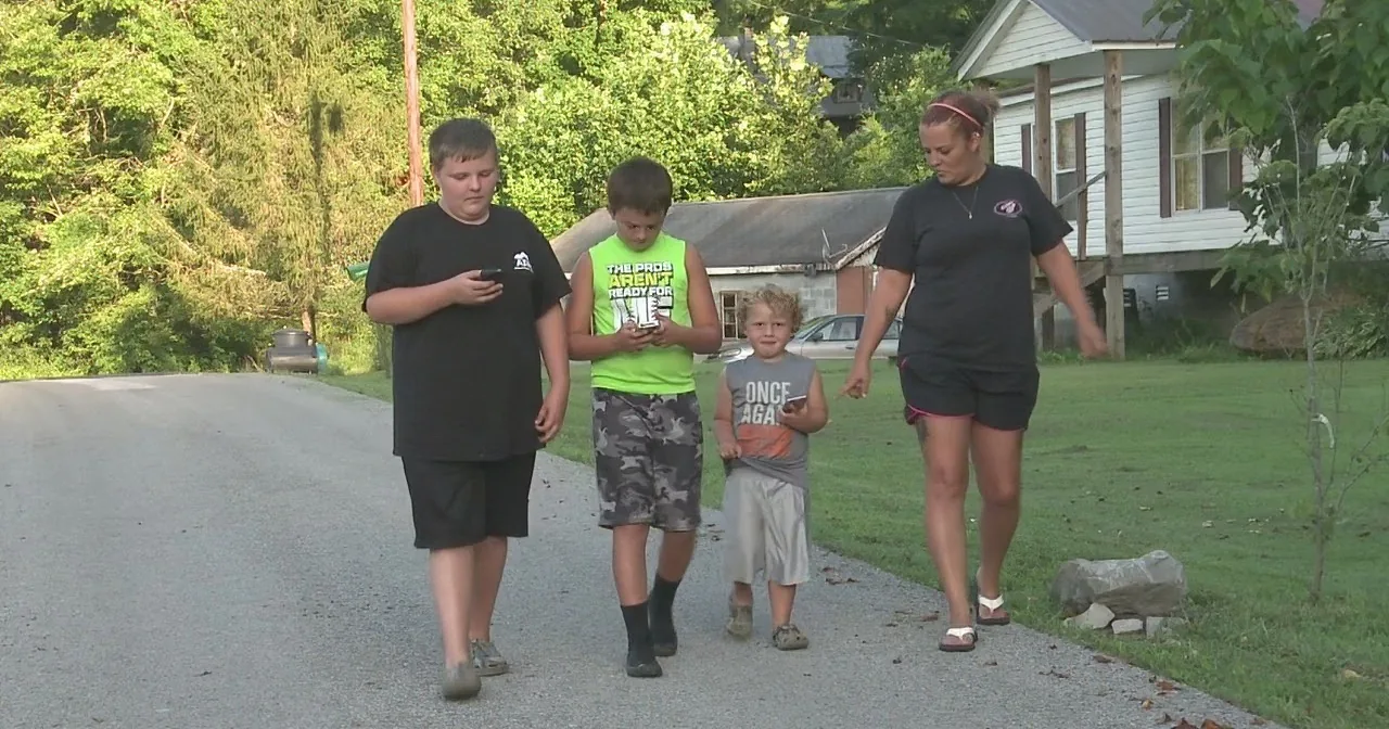 A woman and three children walk on a gravel path near a house and trees.