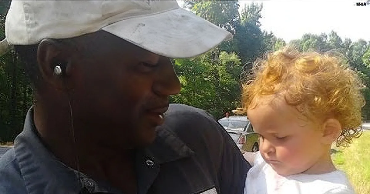 A man wearing a cap and earphones is interacting with a child with curly hair, both outdoors.