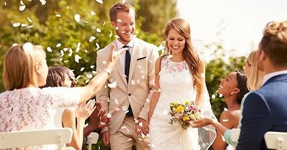 A bride and groom smile amidst falling petals, surrounded by guests in a sunny outdoor setting.