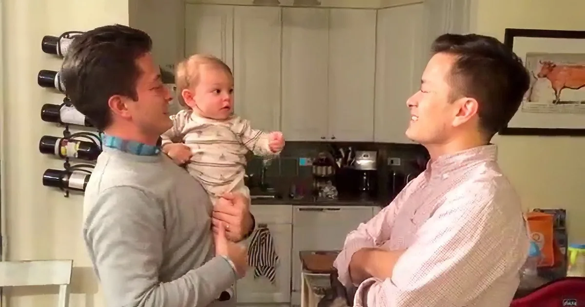 Two men in a kitchen, one holding a baby, both smiling and interacting.