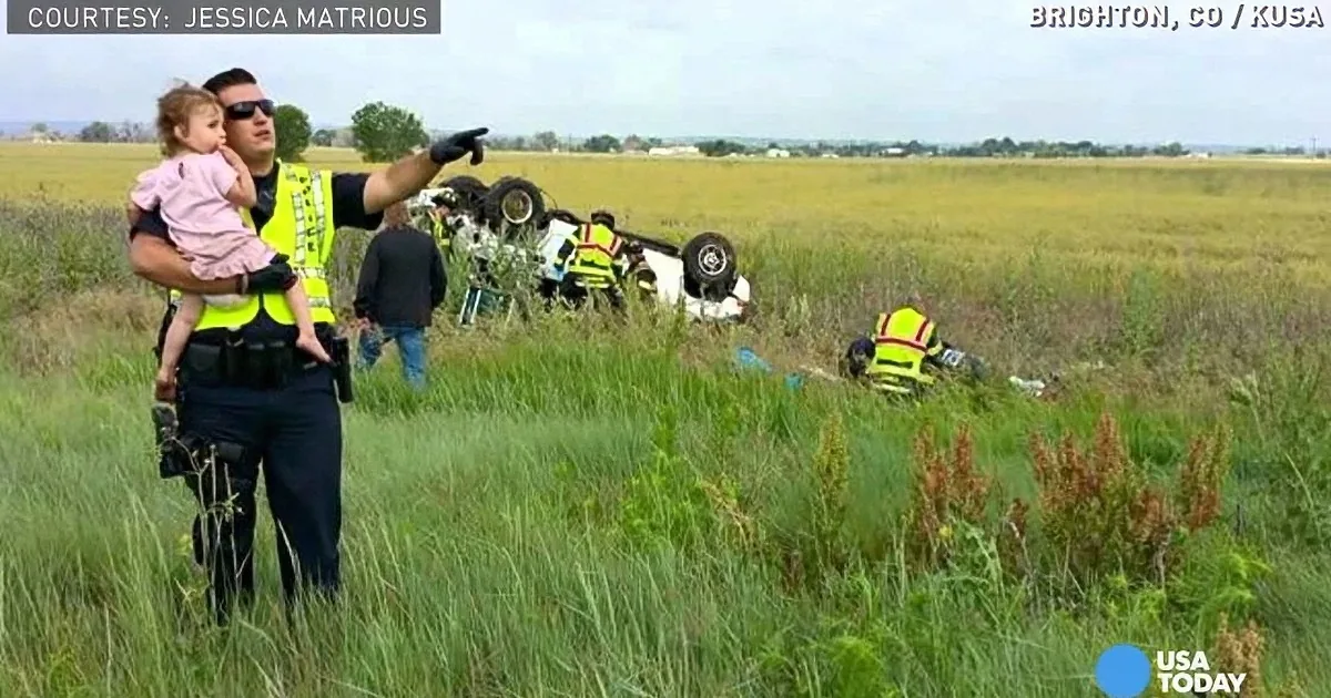 A police officer holds a child while pointing at a flipped car in a grassy field.
