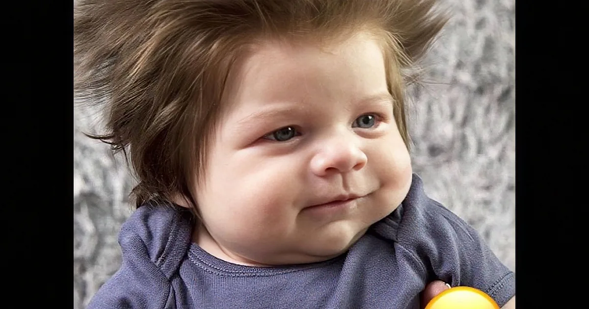 A baby with messy hair and a blue shirt, holding a yellow object.