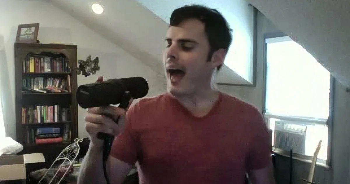 A man in a red shirt holds a hairdryer, singing or speaking with his eyes closed in a room with a bookshelf and window.