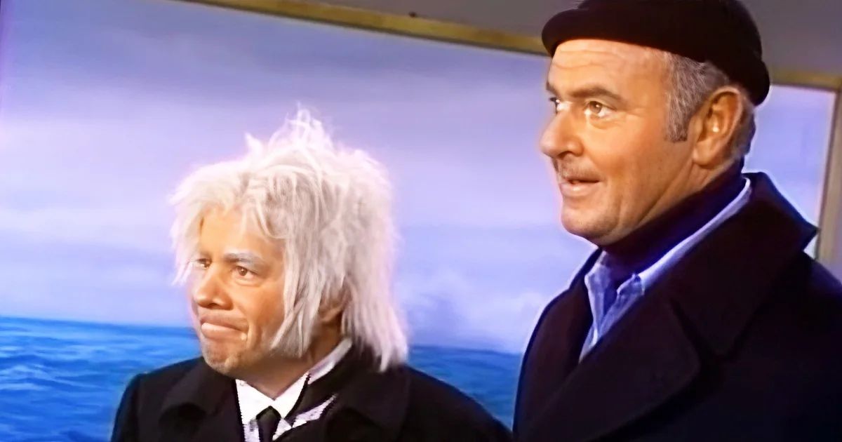 A man with white hair and a black coat stands next to another man in a black beanie and suit, both in front of a scenic...