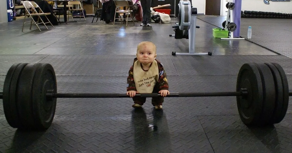 A baby wearing a bib stands between two large weight plates in a gym.