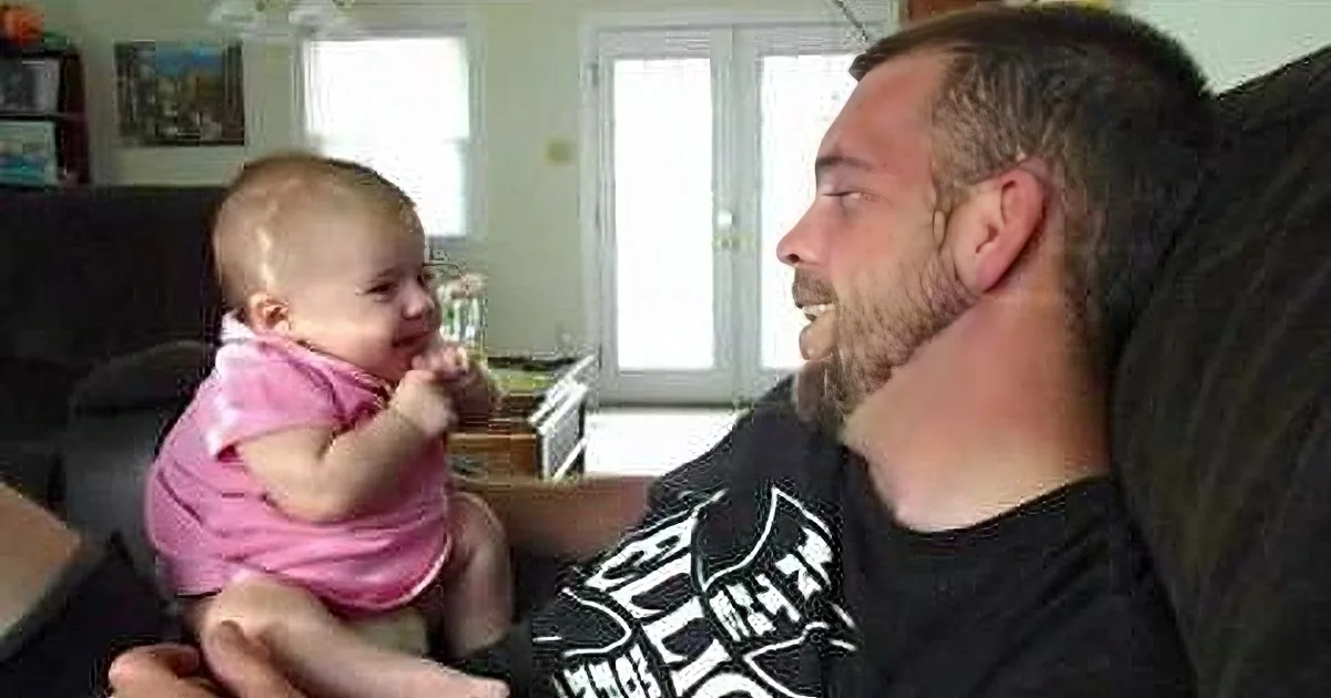 A man and a baby are sitting together, the baby in a pink outfit, both smiling and looking at each other.