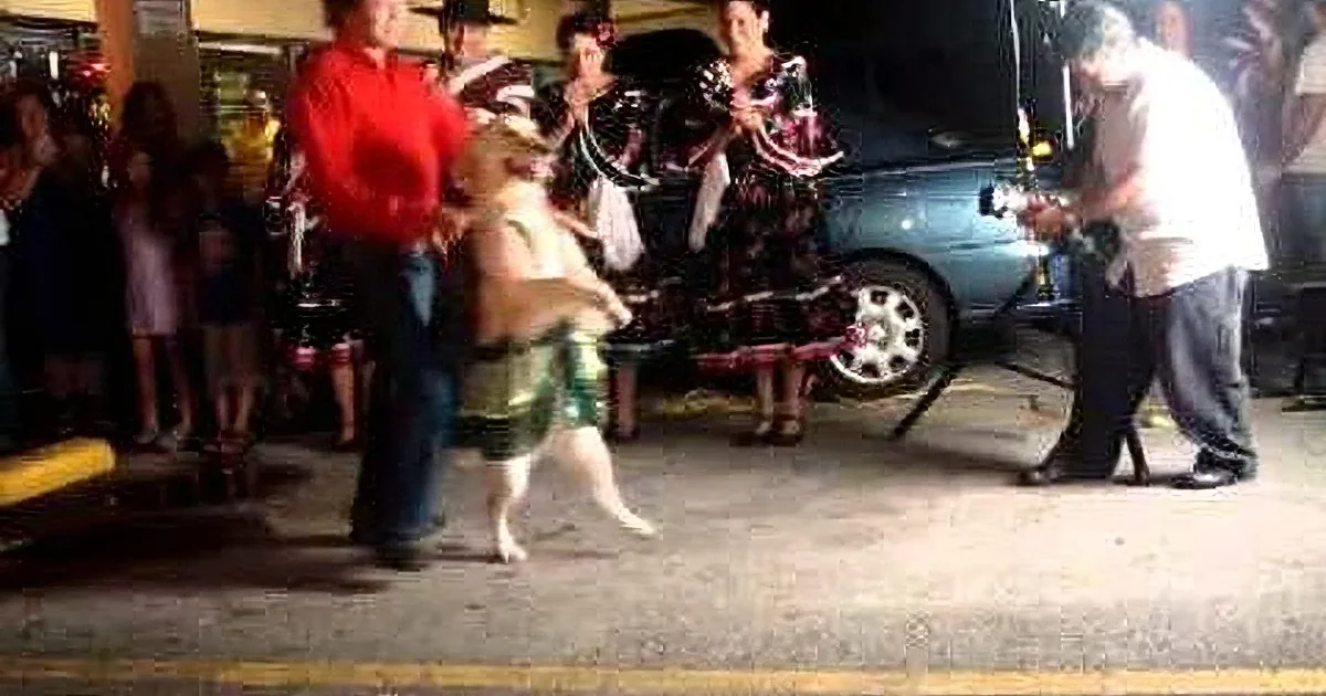 A man in a red shirt and a child in a green skirt are dancing in a parking lot, surrounded by onlookers.