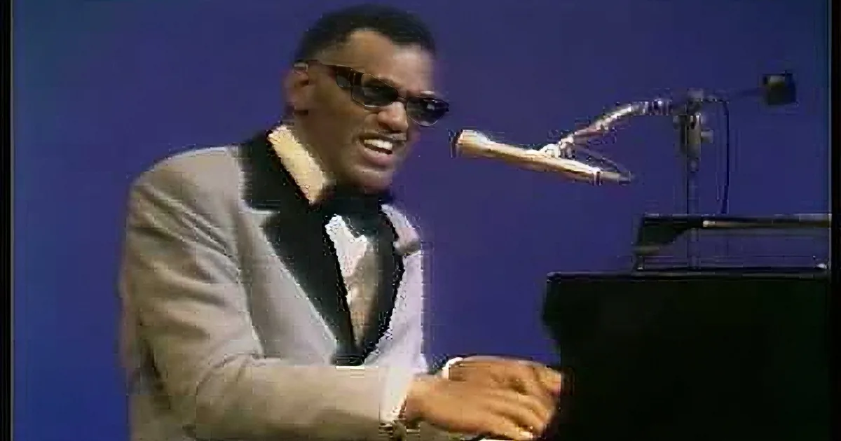 A man in a light suit and sunglasses plays piano on stage with a microphone.