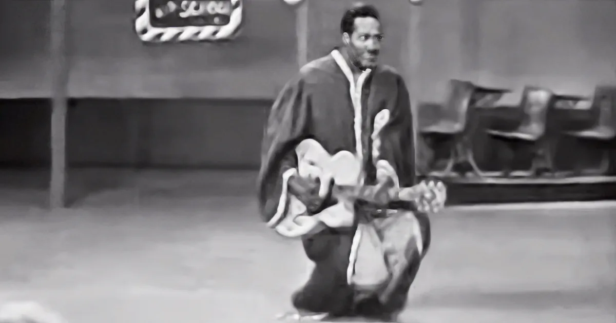 A man in a robe plays guitar on a stage with a sign in the background.