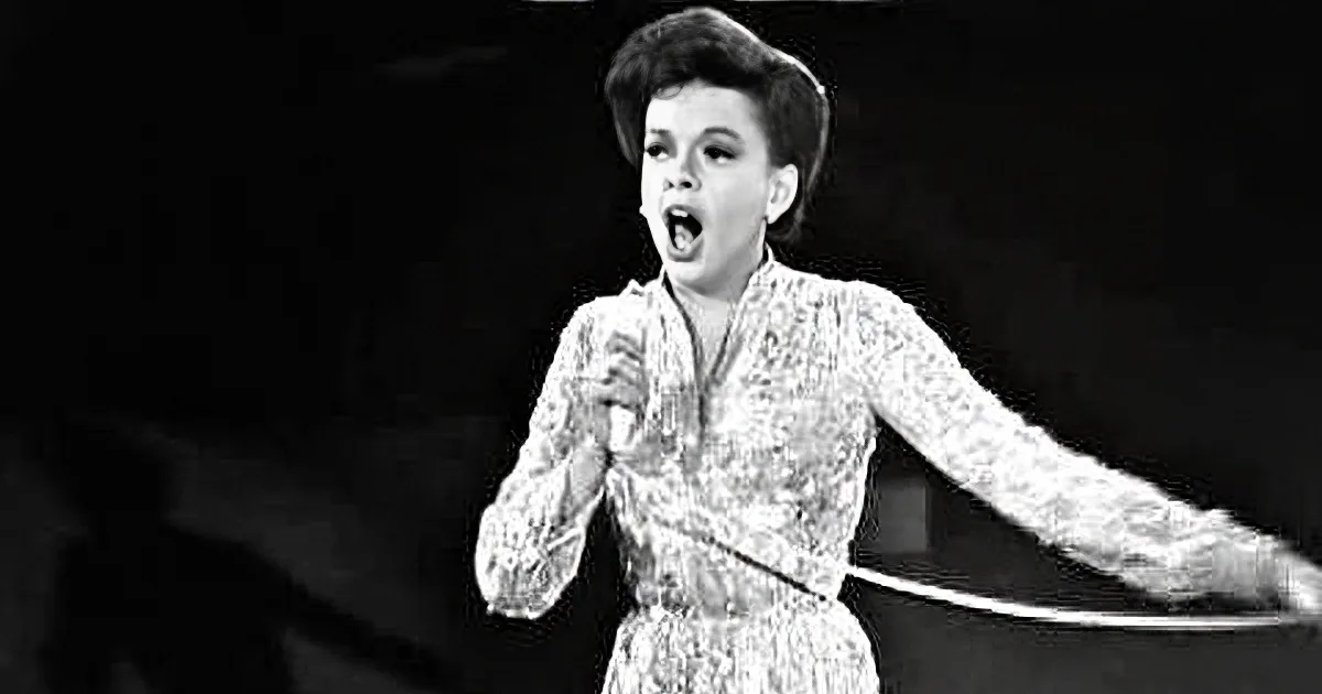 A woman in a patterned dress sings passionately, her arms outstretched, in a black-and-white image.