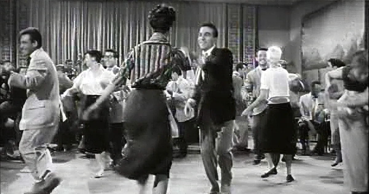 A man in a suit dances with a woman in a striped shirt in a crowded room.