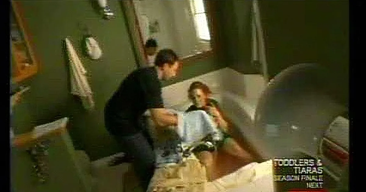 A man leans over a woman lying on a couch in a dimly lit room with green walls.