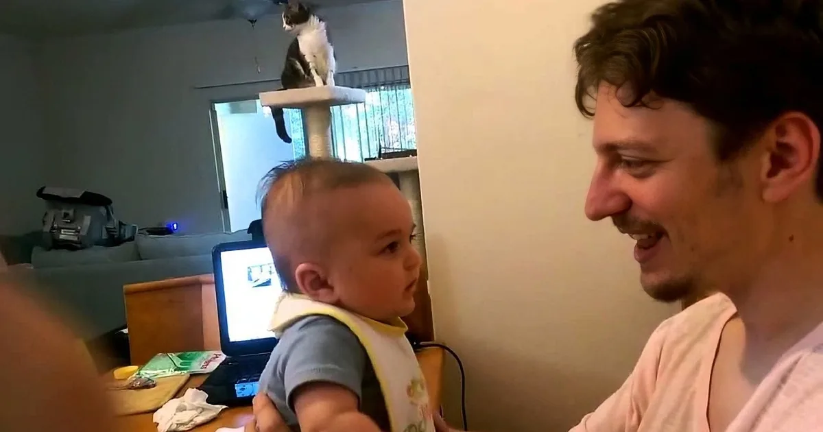 A man holds a baby while smiling, with a cat perched on a scratching post in the background.