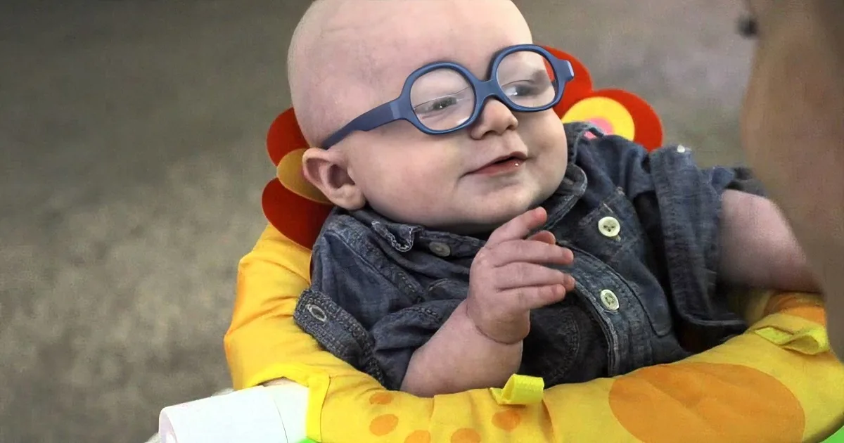 A baby wearing blue glasses sits in a colorful baby bouncer, looking to the side with a curious expression.