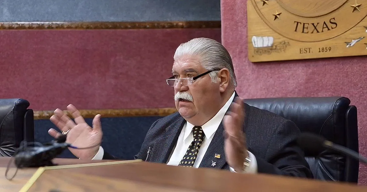 A man in a suit and tie gestures while seated at a desk with a Texas emblem in the background.