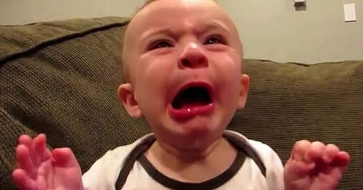 A baby in a white shirt is crying with arms outstretched, sitting on a green couch.