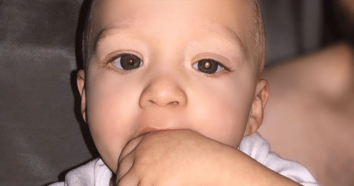 A baby with a hand in its mouth, looking curious and slightly chubby.