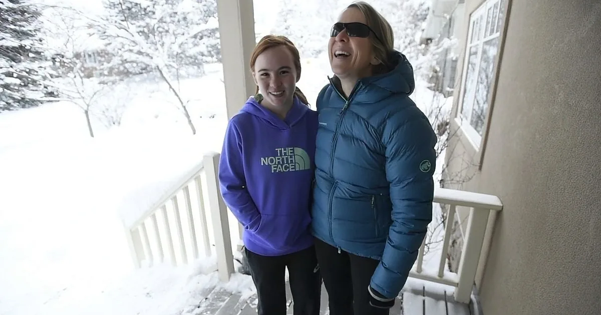 Two people in winter gear smile on a snowy porch, enjoying the cold weather.