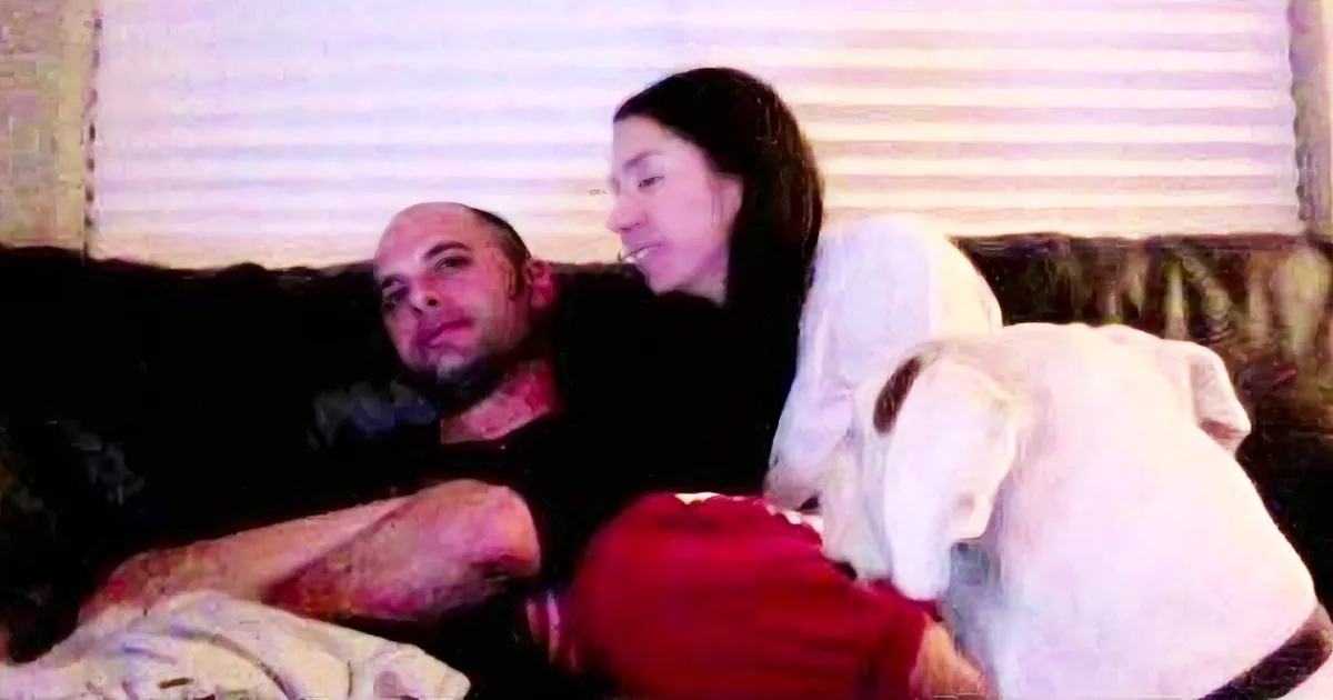 A man and woman sit on a couch with a dog, both smiling and looking at something off-camera.