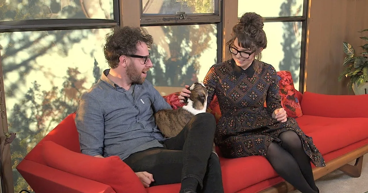 A man and woman sit on a red couch, laughing and petting a dog.