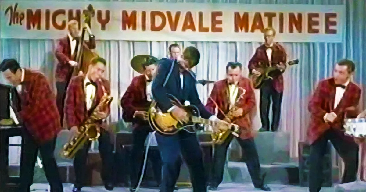 A man in a blue suit plays guitar on stage with a band in red jackets.