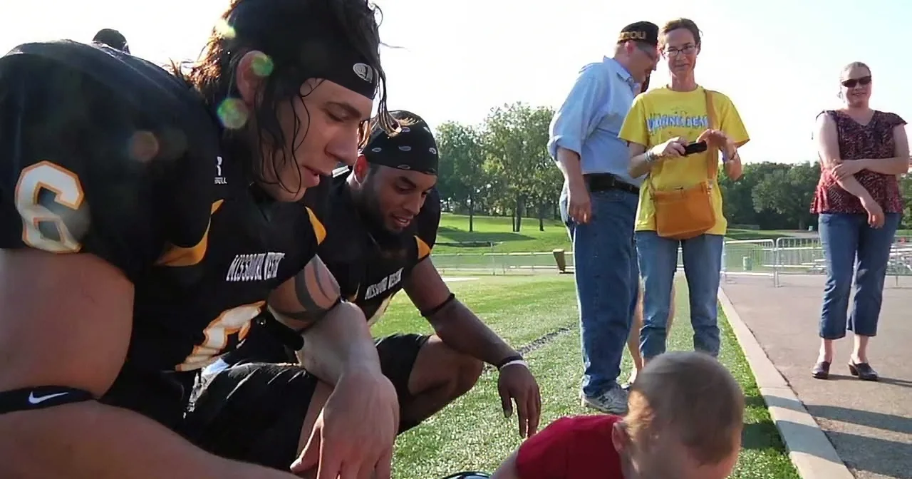Football players in black uniforms interact with a child on a sunny day.