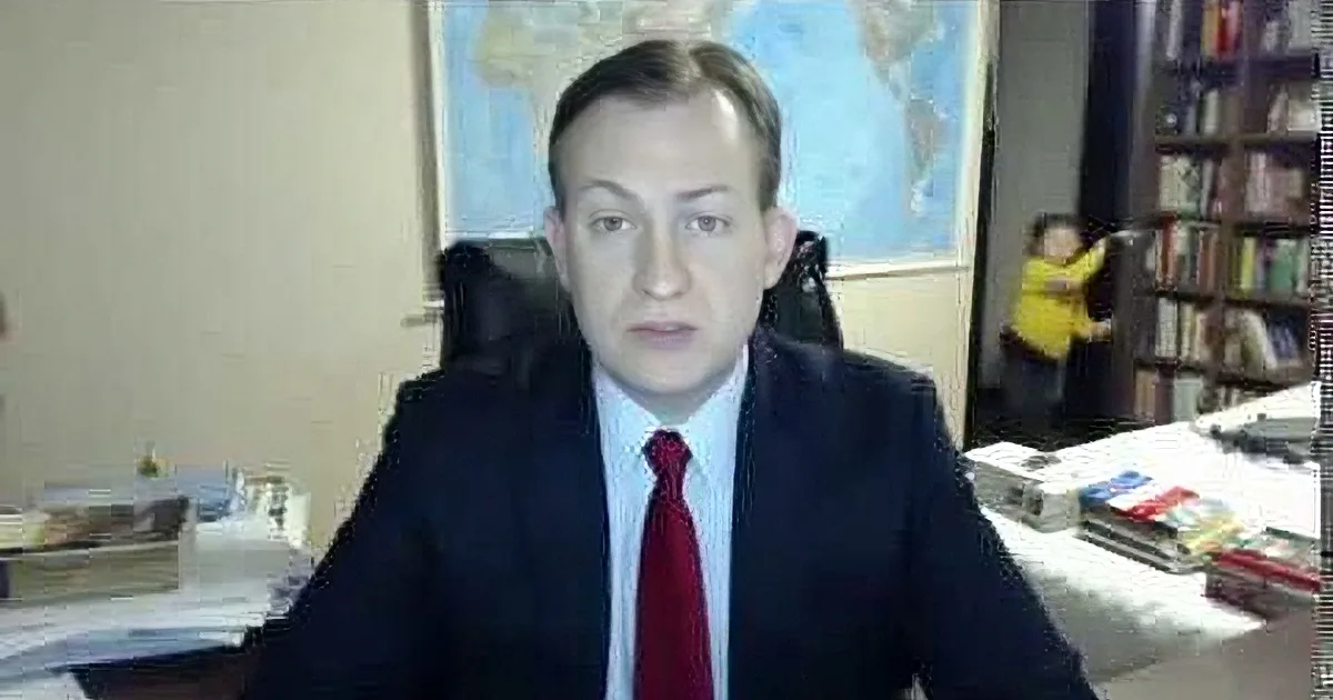A man in a suit and tie sits at a desk, surrounded by books and papers, with a child in the background.