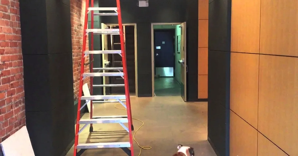 A red ladder leans against a brick wall in a hallway with a door and hallway leading further.