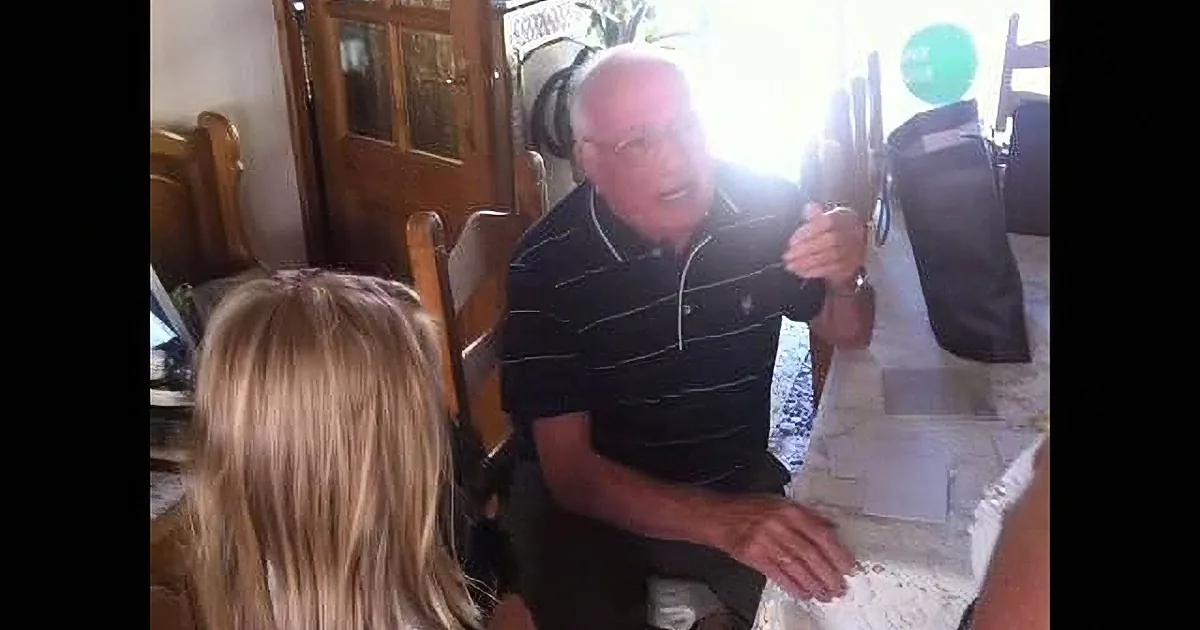 An older man in a striped polo shirt is seated at a table, gesturing with his right hand while speaking.