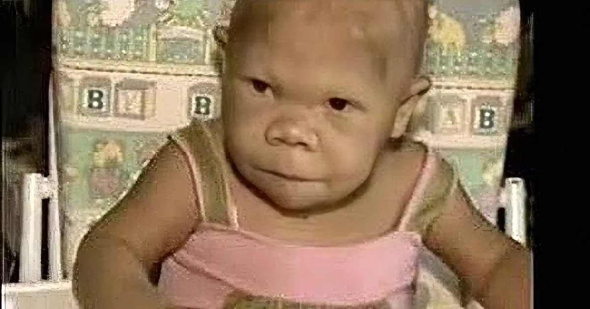 A baby wearing a pink and green outfit sits in a crib with a patterned background.