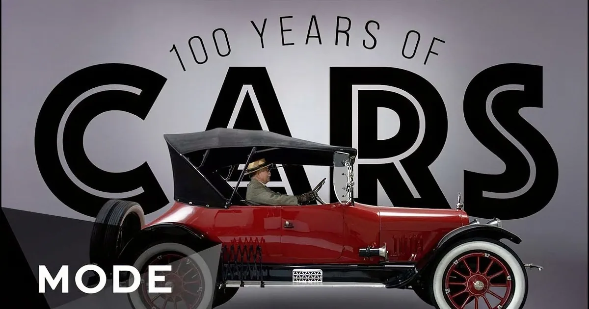 A vintage red car with white-walled tires and a man in a hat driving it.