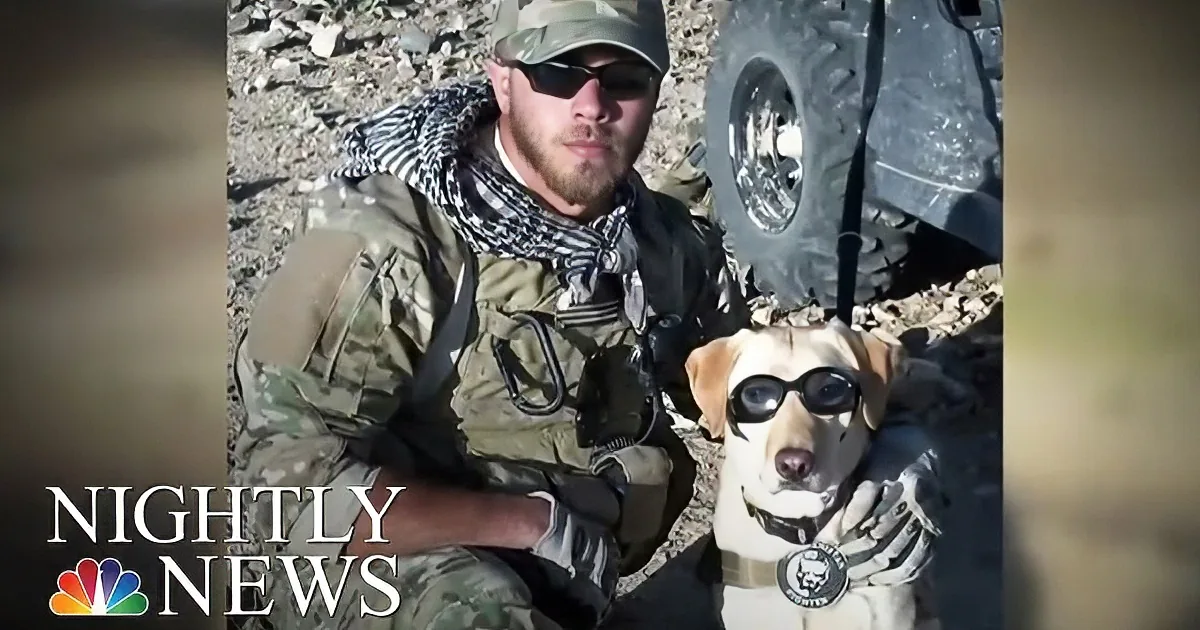 A soldier in camouflage holds a dog wearing goggles and a harness.