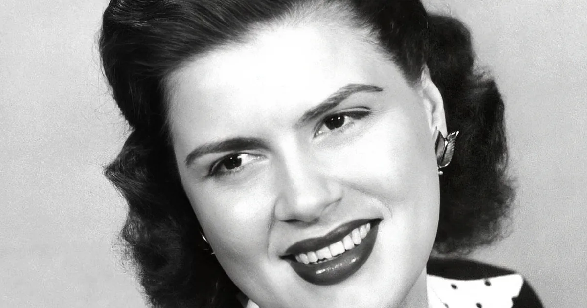 A woman with dark, styled hair, wearing a polka-dotted outfit and a butterfly earring, smiles warmly.