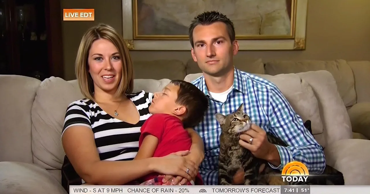 A family of three sits on a couch, smiling, with a cat on the man's lap.