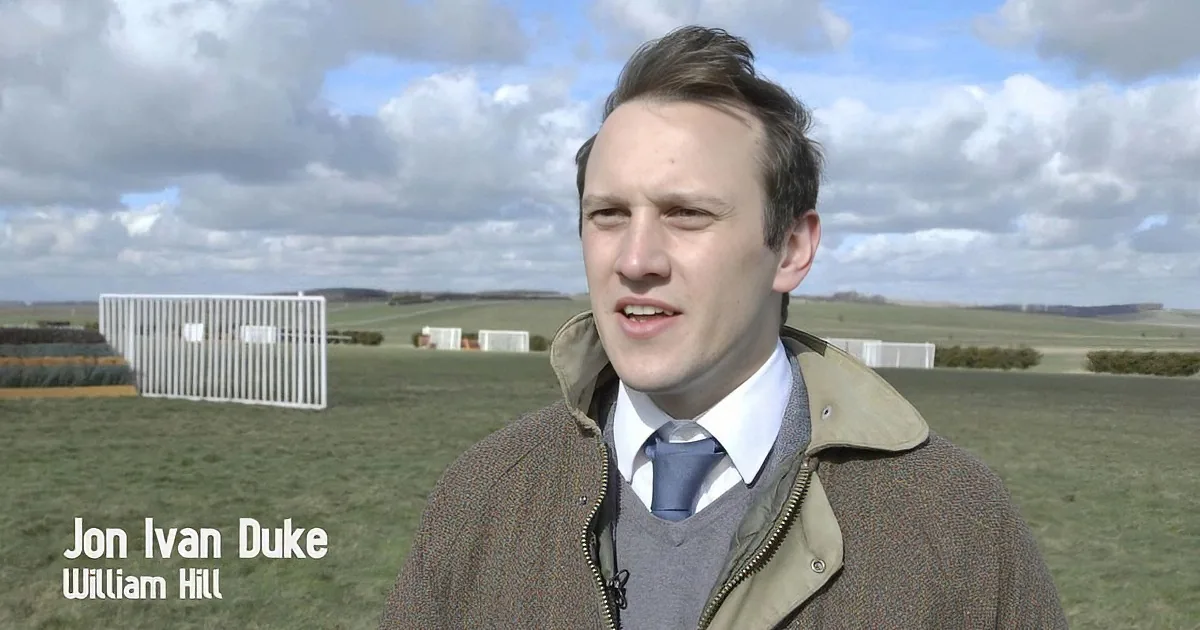 A man in a brown jacket and blue tie stands in a grassy field with a cloudy sky and a white structure in the background.