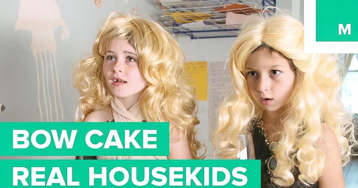 Two children with blonde wigs, one wearing a black dress, in a room with a wall decoration.