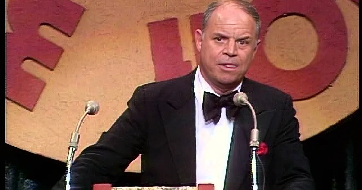 A man in a tuxedo and bow tie stands at a podium with microphones, speaking in front of a red and yellow backdrop.