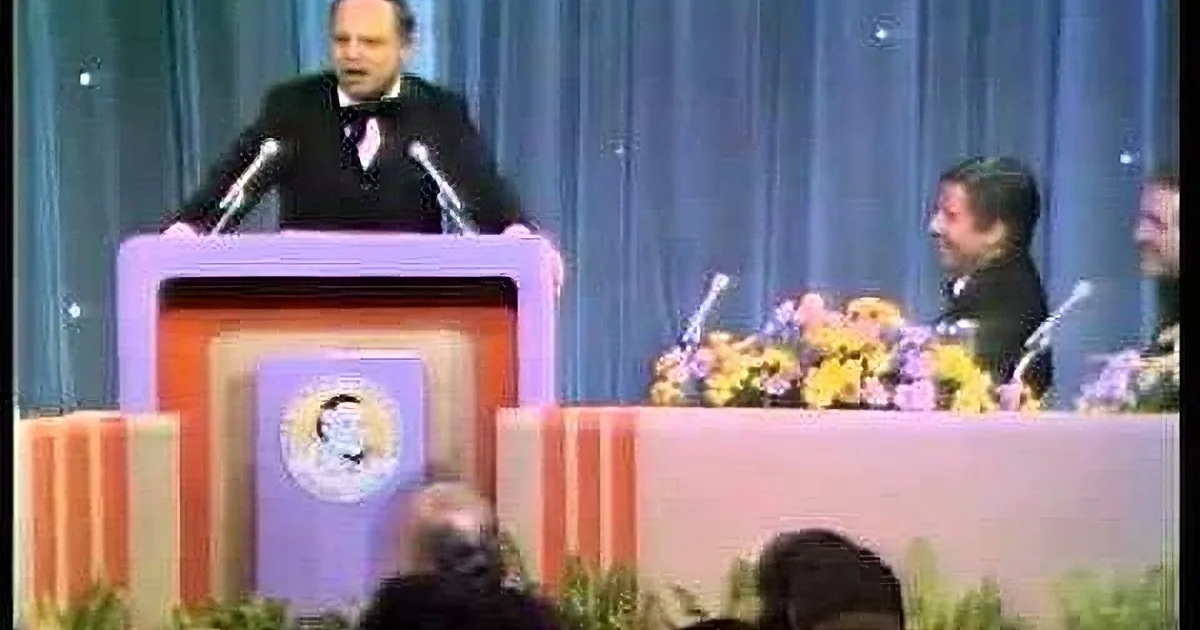 A man in a tuxedo stands at a podium with a microphone, addressing an audience.