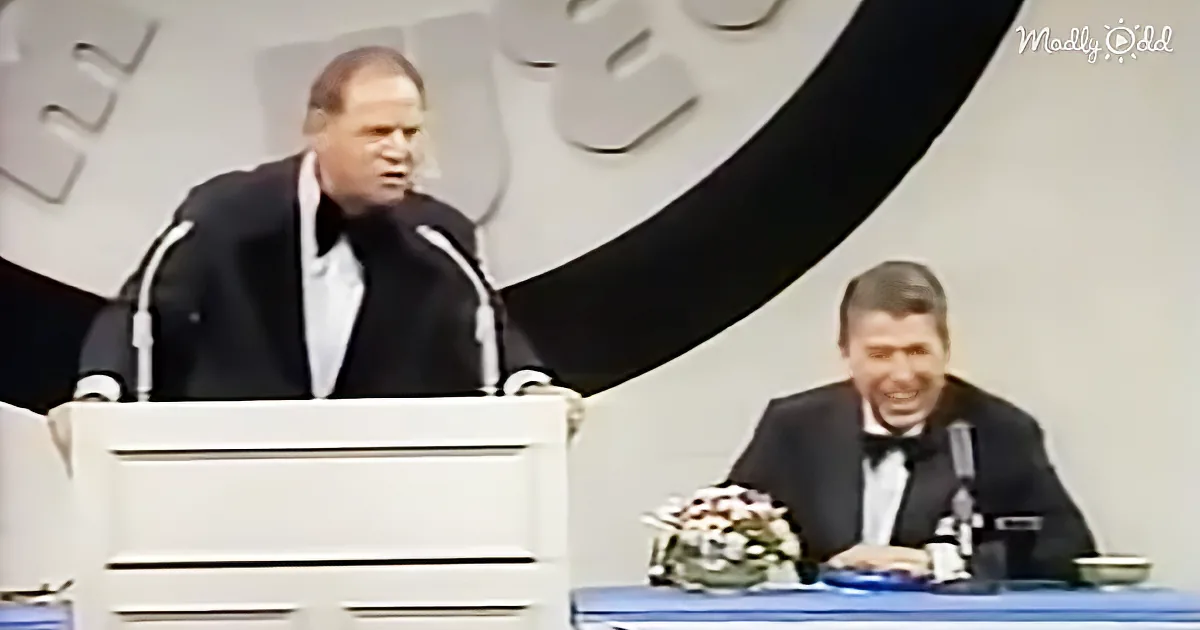 A man in a suit stands at a podium, speaking, while another man in a tuxedo sits at a table with a flower arrangement.