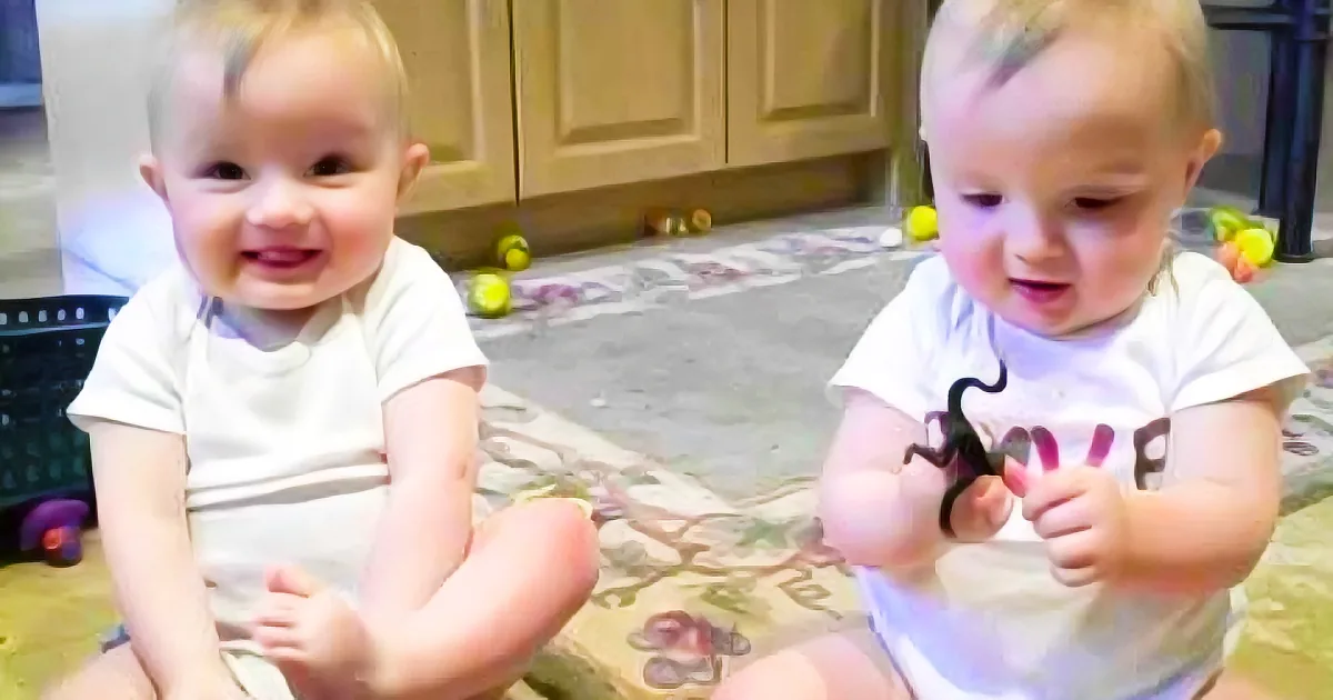 Two babies, one smiling, both playing with toys on a patterned rug in a kitchen.