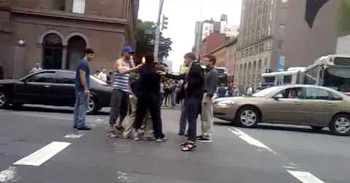 A group of people stands in a city street, with a man in a blue shirt and another in a striped shirt prominently featured.