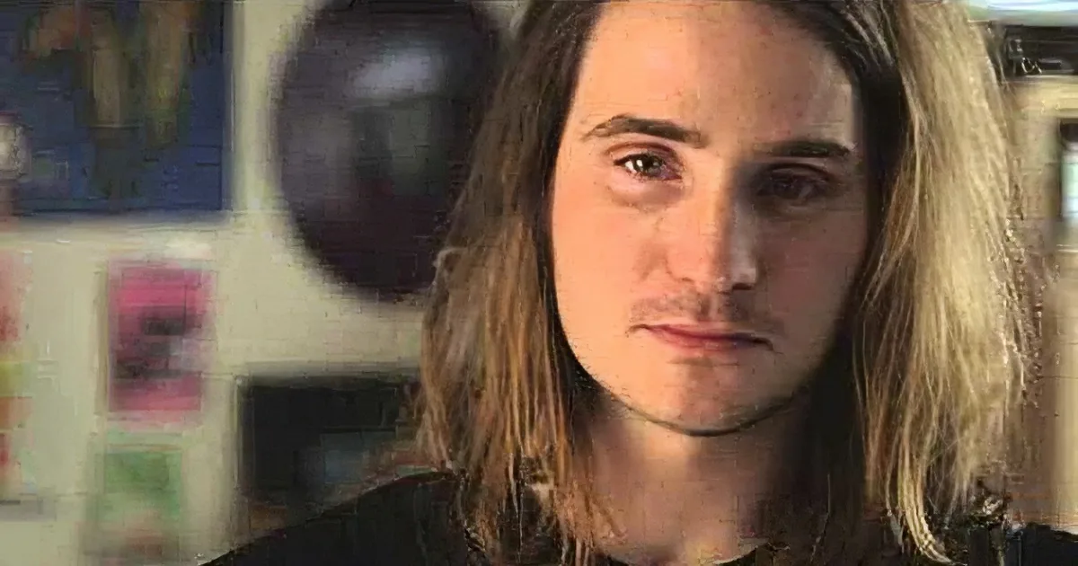 A person with long hair, wearing a black shirt, in a room with various posters and objects in the background.