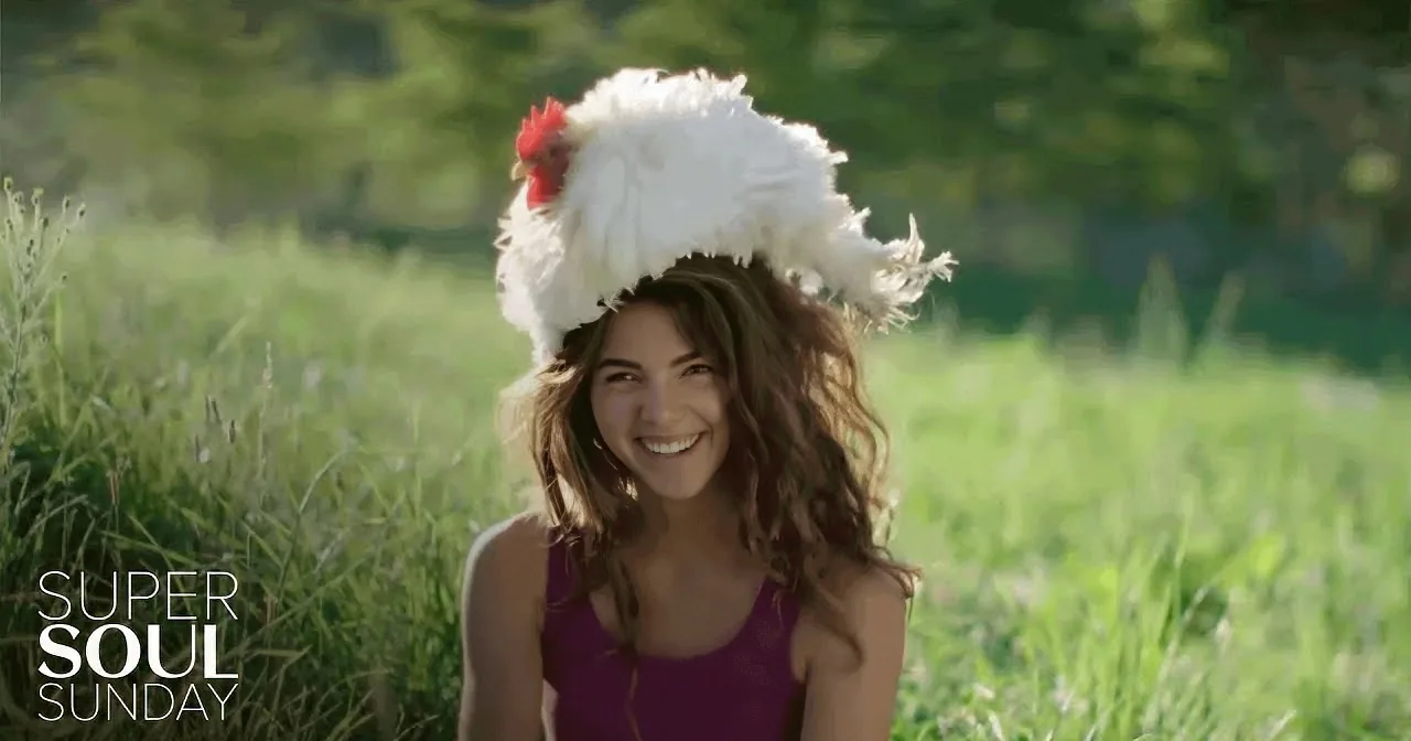 A woman in a field wears a large white chicken hat, smiling.