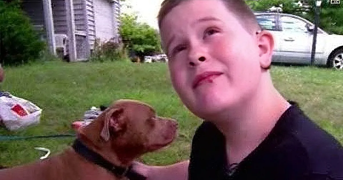 A young boy and a brown dog are in a grassy yard, with a house and car in the background.