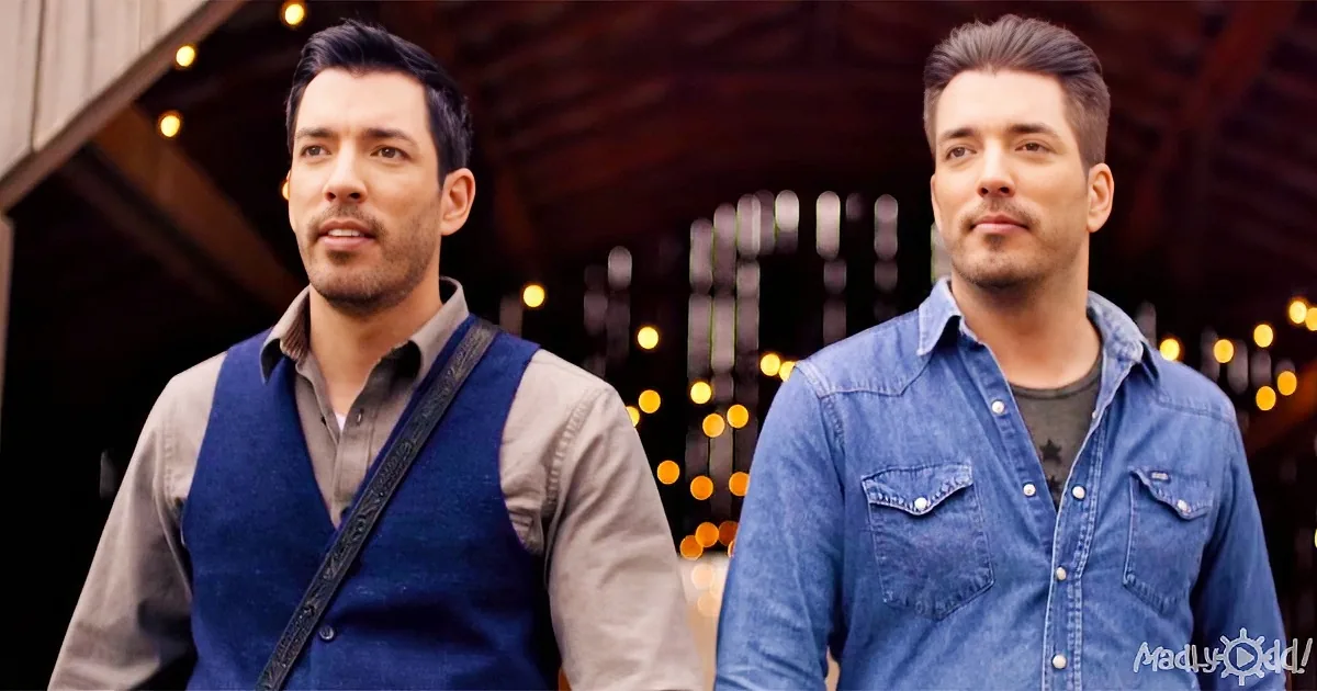 Two men in casual attire stand side by side, one in a vest and the other in a denim shirt, against a backdrop of warm lights.